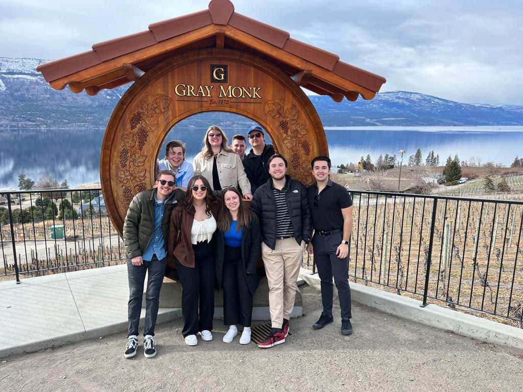 Group of people posing in front of a wooden archway with "Gray Monk" inscription, overlooking a scenic lake and vineyard landscape, representing a visit to local attractions near Cozy Coffee in Kelowna, BC.