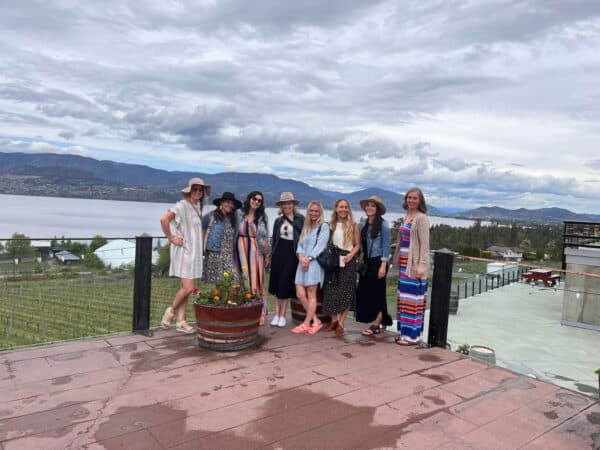 Group of women posing together on a patio with a scenic view of Okanagan Lake and vineyards, highlighting a social experience related to tours offered by Carpe Diem Tours.