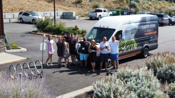 Group of tourists posing joyfully in front of a Carpe Diem Tours shuttle van, with a parking lot and greenery in the background, highlighting travel experiences in the Okanagan area.
